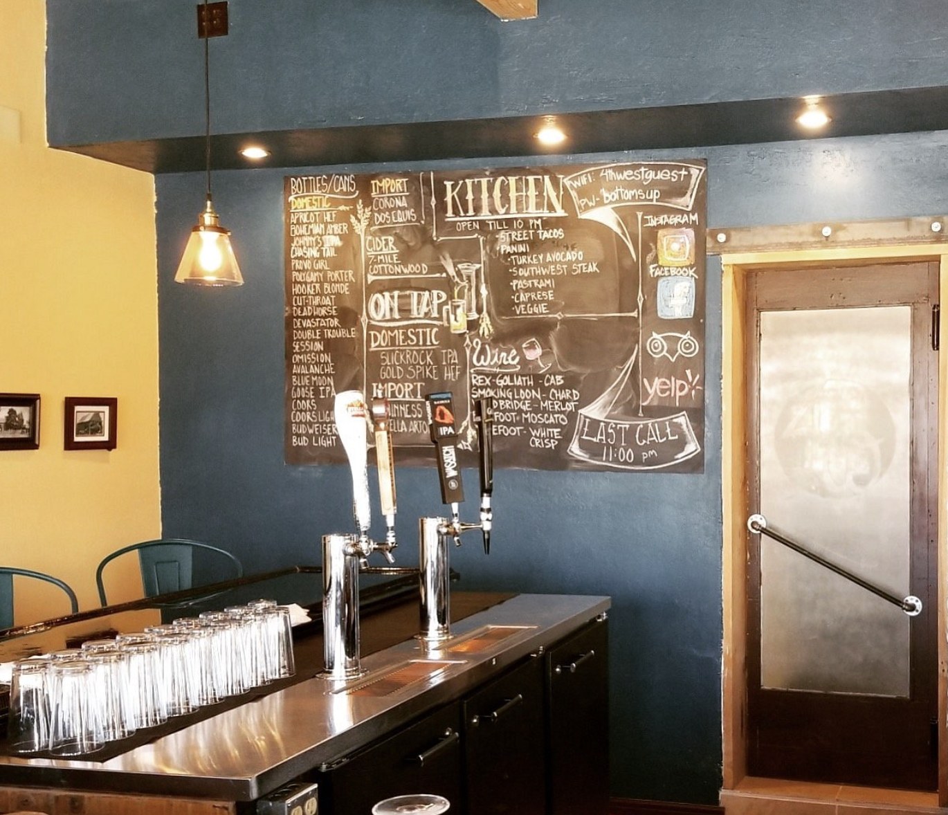 A chalkboard menu listing street tacos, sandwiches, and wine inside 4th West Pub in Escalante with beer taps in the foreground