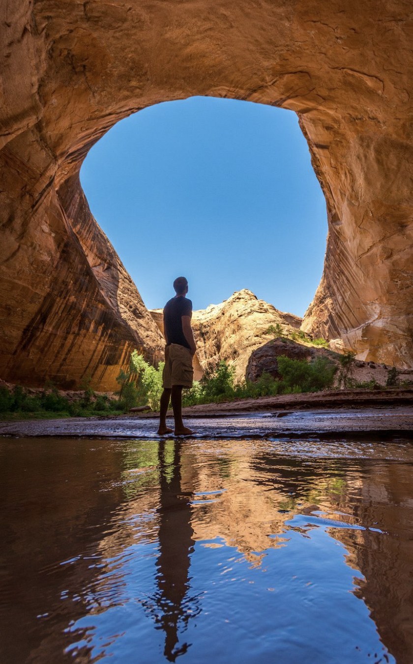A hiker standing in a shallow pool beneath a sandstone arch with the sky reflected in the water