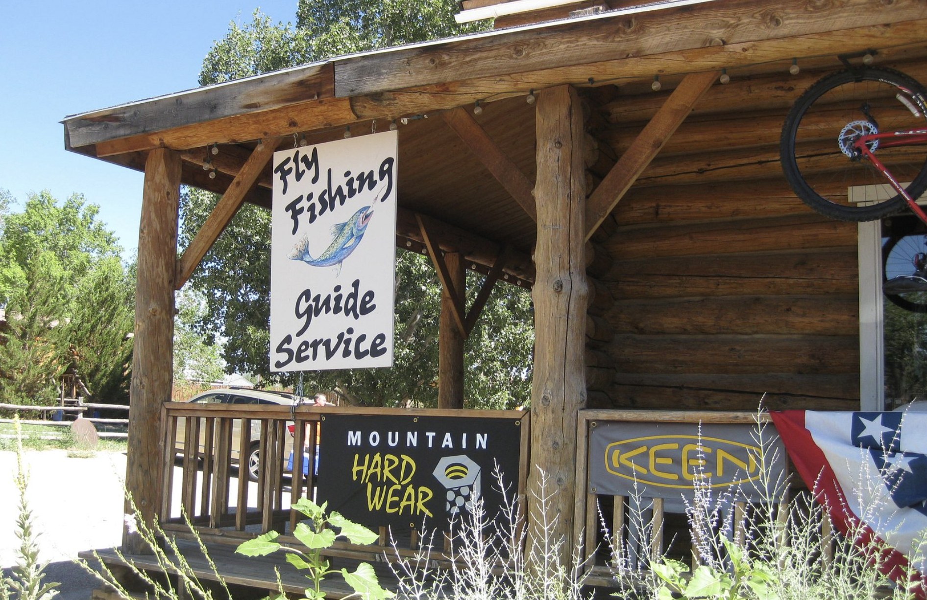A Fly Fishing Guide Service sign hanging on a log cabin porch with outdoor gear brand banners in Escalante