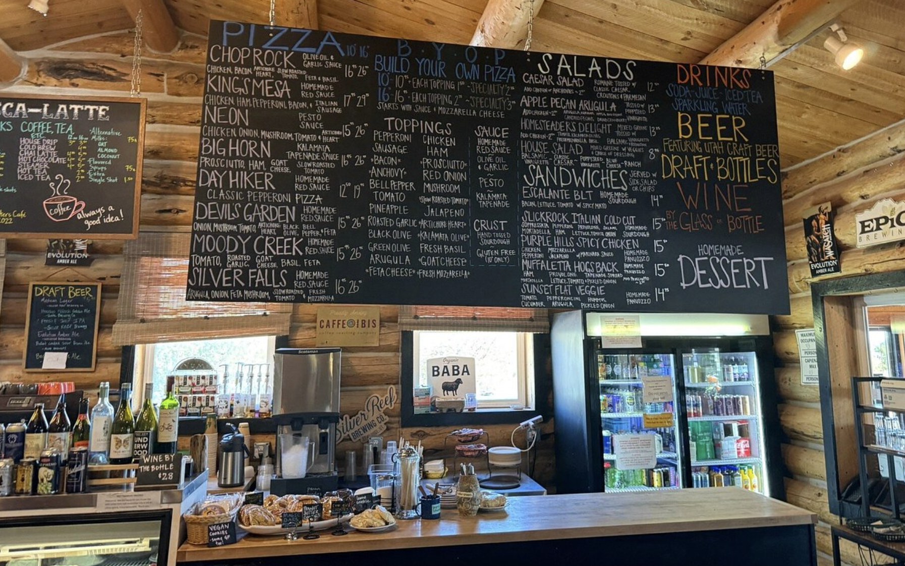 A large chalkboard menu listing pizzas, salads, sandwiches, and drinks inside a cafe in Escalante