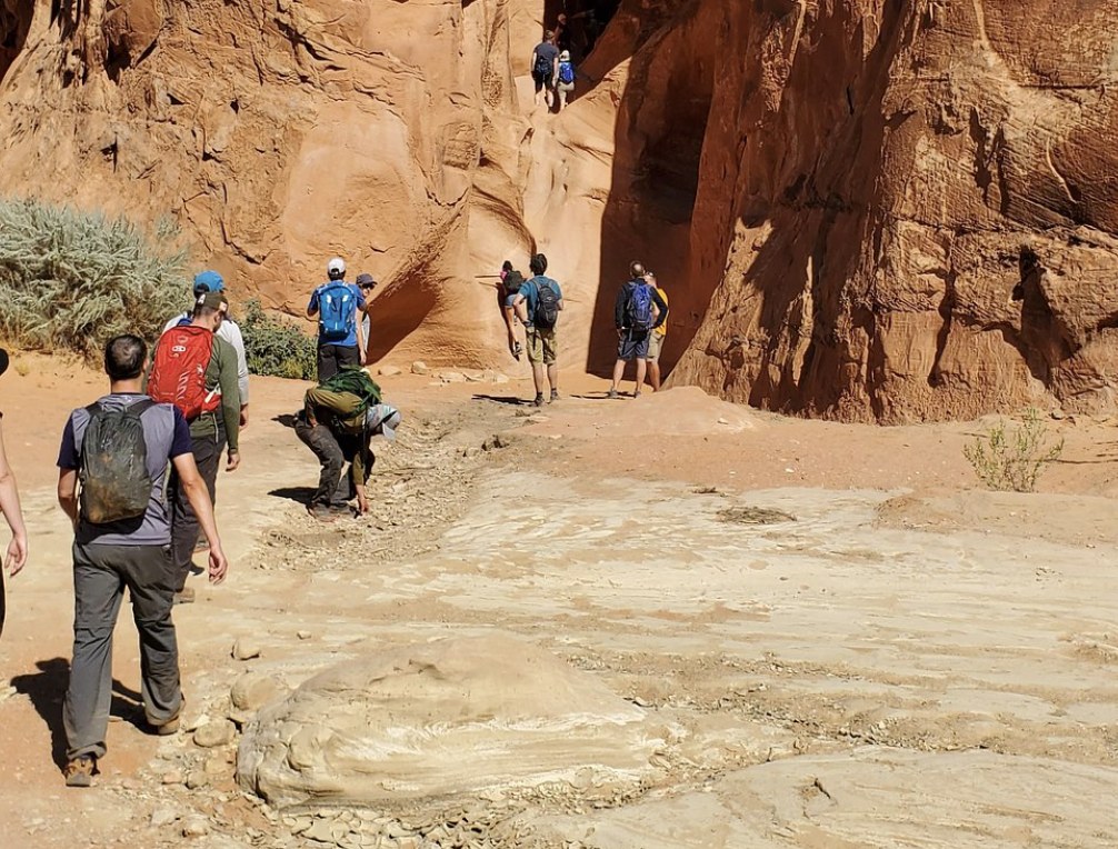 A group of hikers with backpacks walking toward the entrance of a narrow slot canyon near Escalante