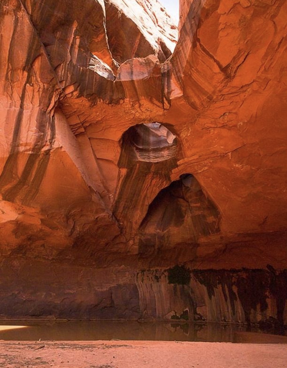 Round skylight openings in a red sandstone alcove above a still pool of water