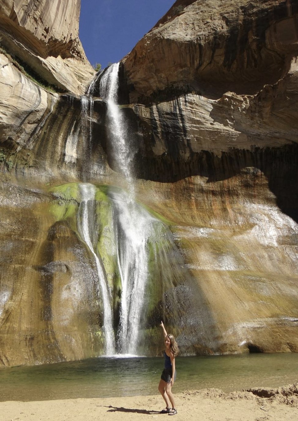 A woman standing at the base of Lower Calf Creek Falls, pointing up at the waterfall for scale